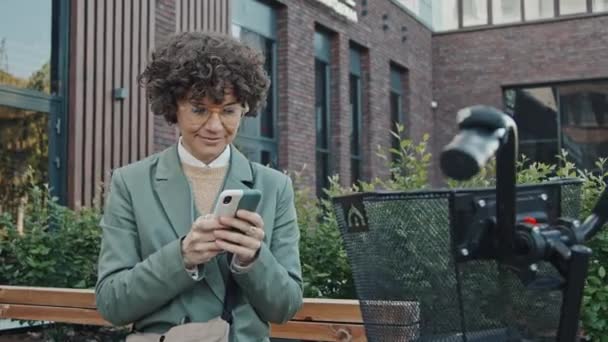 スマートフォンで何かをテキストするベンチに座っているカーリーヘアの現代白人女性の中型肖像画 — ストック動画
