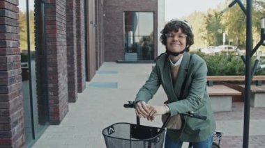 Kameraya gülümseyen bisikletiyle dışarıda duran gözlüklü ve kasklı şık giyinen neşeli beyaz iş kadınının orta boy portresi.