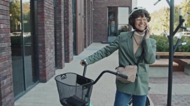 Kafkaslı, şık giyinen, kasklı, bisikletle sokakta telefonla konuşan neşeli beyaz bir kadının orta boy fotoğrafı.