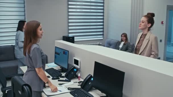 現代病院のチェックインデスクで患者と一緒に働く陽気な若い白人女性 中長いショット — ストック動画