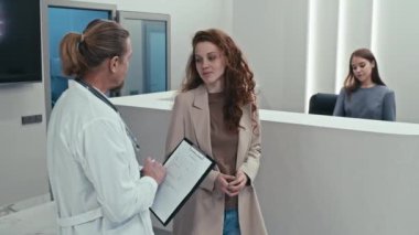 Olgun erkek doktor ve genç bayan hasta modern hastane lobisinde durmuş sohbet ediyorlar.