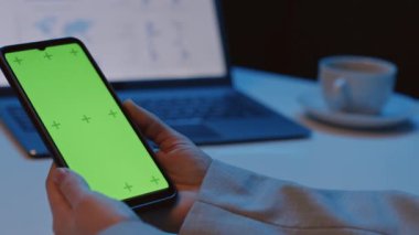 Gece vakti solda, elinde yeşil ekran olan akıllı telefonu tutan bir kadın var.