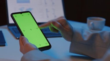 Masada oturan, işaret parmağını hareket ettiren, gece vakti yeşil ekranlı, akıllı telefonu hareket ettiren, tanınmayan bir kadının ahır görüntüsü.