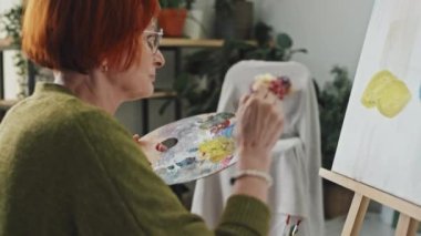 Yeşil hırkalı, orta yaşlı bir kadının resim çekimi. Şövalenin önünde oturmuş yağlı boya ve fırçayla resim yapıyor.