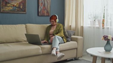 Sol tarafta oturan, kırmızı saçlı, evde oturan, kulaklık takan ve dizüstü bilgisayarına bakan yaşlı bir kadın resmi var.