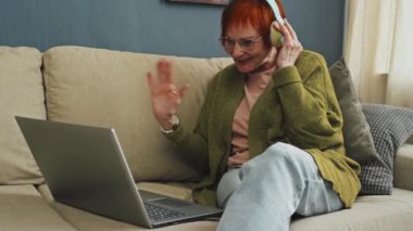 Kulaklık takmış, gözlüklü, gülümseyen, dizüstü bilgisayarlı ve video görüşmesi sırasında ailesiyle konuşan yaşlı bir kadın.