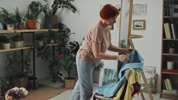 着るものを選択する居心地の良いスタジオで鏡の前に立っている赤い髪のシニア女性のミディアムフルショット — ストック動画