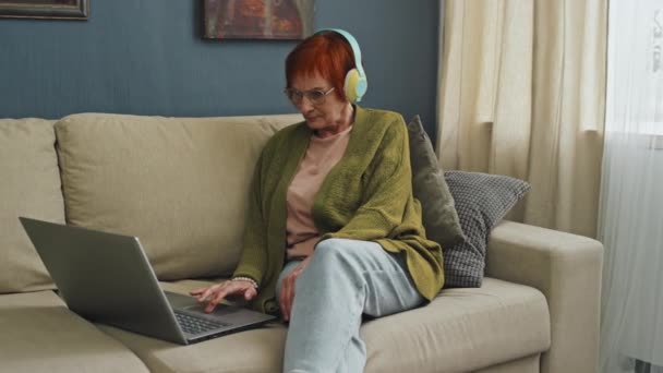 ラップトップを見ているアパートでソファーに座っている赤い髪のシニアの女性の左中型ショット — ストック動画