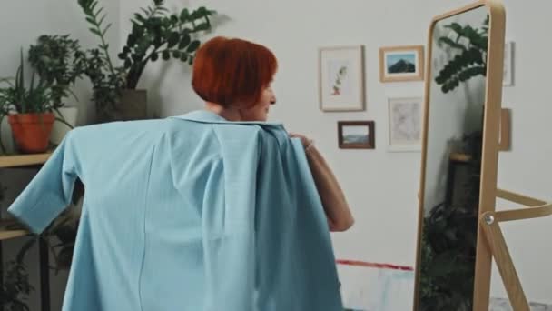 日中の鏡の前で青いジャケットを試しているスタジオに立っている笑顔のシニア女性のメディアショット — ストック動画