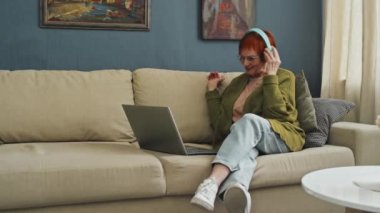 Kanepede dizüstü bilgisayarıyla oturmuş video görüşmesi sırasında akrabalarına merhaba diyen neşeli son sınıf kadınlarının tam görüntüsü.