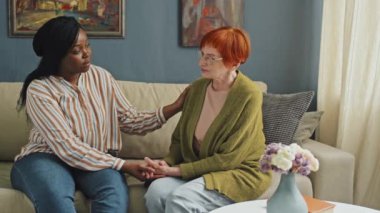 Orta ölçekli sol çekte, yanında oturan ve hayal kırıklığına uğramış yaşlı bir kadının omzuna ve eline dokunduğu Afro-Amerikan ev bakımı asistanı.