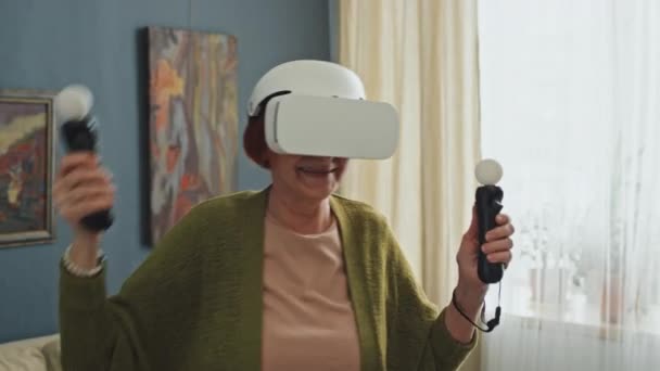 戴Ar护目镜和手持运动传感器在公寓中央玩游戏的老年妇女的中等特写镜头 — 图库视频影像