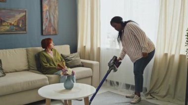 Oturma odasındaki koltukta oturan son sınıf beyaz kadın ve yerleri süpüren genç Afrikalı Amerikalı kadın.