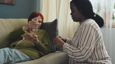 Orta boy beyaz bir kadın oturma odasında kanepede uzanıyor ve Afro-Amerikan ev bakımı asistanı yanında oturmuş bir bardak su ve hap veriyor.