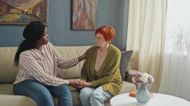 パンニングは アフリカ系アメリカ人の若い女性のミディアムフルショットを残し 昼間のアパートで赤髪の問題を抱えたシニアレディをサポートしました — ストック動画