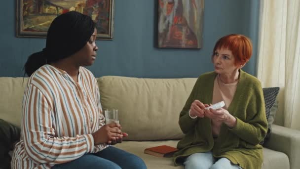 アフリカ系アメリカ人のホームケアアシスタントのミディアムショットは リビングルームのソファーに座って 赤髪のシニア女性に水を与える — ストック動画