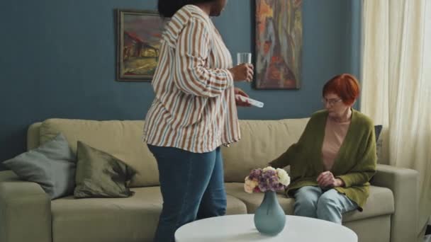 女性のアフリカ系アメリカ人のホームケアアシスタントのフルショットは 昼間のリビングルームにソファに座って赤髪のシニア女性に丸薬を与えます — ストック動画