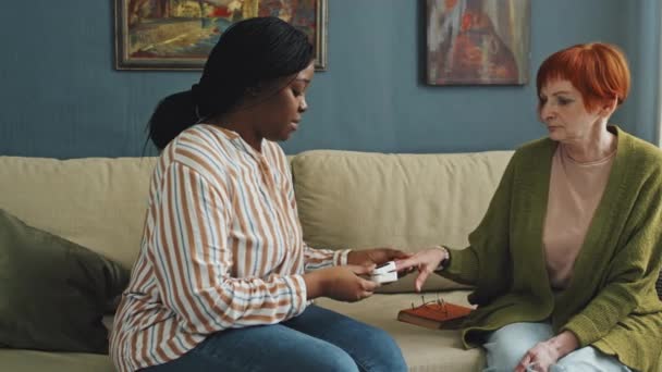アフリカ系アメリカ人女性がコーカサス人のシニア女性とソファーに座り 昼間はリビングルームに指のセンサーを手に入れました — ストック動画