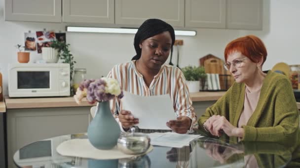 拍下了一张非洲裔美国女人的右半张照片 她一边看医生处方 一边和坐在厨房餐桌旁的白人老年妇女交谈 — 图库视频影像