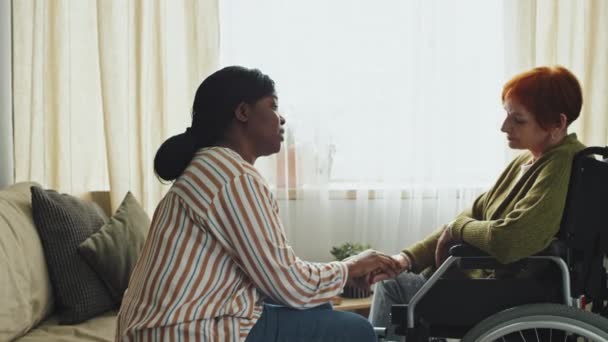 坐在轮椅上的老年妇女和非洲裔美国人家庭护理助理在白天让她平静下来 并将她的手伸进客厅的中间镜头 — 图库视频影像