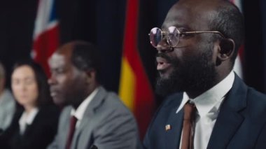 Uluslararası kriz zirvesinde duygusal konuşmalar yapan Afro-Amerikan erkek bakanı, çok ırklı meslektaşları dinleyen ve arka planda çeşitli devlet bayrakları bulunan orta ölçekli seçici fotoğraf.