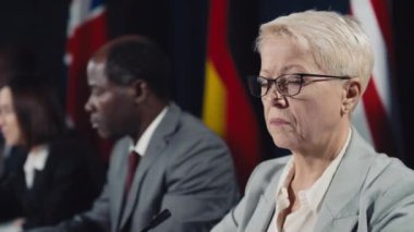 Resmi takım elbiseli, gözlüklü, çeşitli meslektaşları ve bayraklarla uluslararası konferansta oturan beyaz bir kadının orta boy yakın çekimi. Sonra başını çevirip sert bir suratla kameraya bakıyor.