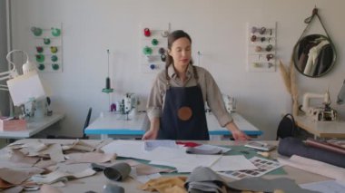 Kafkas desenli önlüklü güzel bir kadının orta boy portresi atölyede deri civata ve moda çizimleriyle duruyor, kameraya bakıyor ve gülümsüyor.