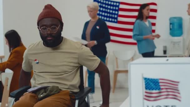 中年坐在轮椅上的非洲裔美国人 戴着便帽和眼镜 骑着马走到投票站印有美国国旗标志的投票箱前 抛出他的公告 然后离开了 — 图库视频影像