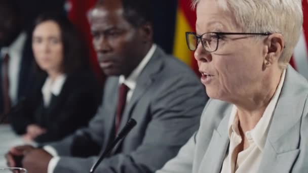 在国际危机峰会上 愤怒的中年金发白人女政治家在国际危机峰会上讲话 不同的国家元首聆听 背景模糊的各种旗帜的特写选择性镜头 — 图库视频影像