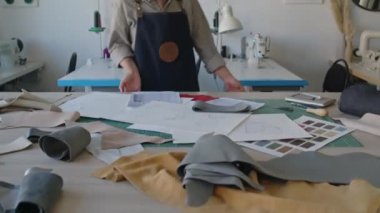 Güzel bayan atölye sahibinin, deri rulo ve ürün çizimleriyle masaya doğru yürüdüğü, kameraya bakıp gülümseyen orta ölçekli yakın çekim portresi.