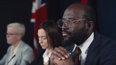 Resmi takım elbiseli, kravat ve gözlüklü sakin ve kendinden emin Afrikalı Amerikalı erkek politikacının hükümet toplantısında otururken çekilmiş yakın plan portresi. Beyaz kadın bakan konuşuyor.
