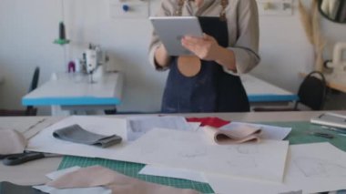 Deri ve ürün çizimleriyle atölyedeki kesim masasında duran, tablet bilgisayar kullanan, kameraya bakan ve gülümseyen kadın moda tasarımcısının yakın çekim eğik portresi.