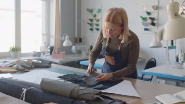 Orta yaşlı sarışın, beyaz önlüklü kadın zanaatkarın atölyede masasında dikilip makasla dikkatlice deri keserken.