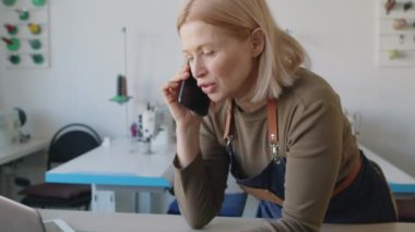 Orta yaşlı, önlüklü, deri atölyeli, kesim masasına yaslanmış, akıllı telefondan konuşan ve dizüstü bilgisayarla çalışan sarışın kadının orta ölçekli yakın çekimi.
