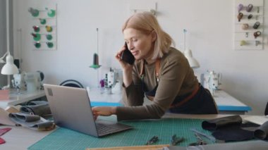Sarışın, orta yaşlı, beyaz önlüklü bir kadının deri atölyesinde masa keserken, dikiş makinelerinin önünde cep telefonuyla konuşurken ve dizüstü bilgisayarına not alırken orta boy bir fotoğraf.