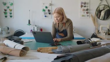 Atölyede duran, deri ve süet cıvataları kontrol eden ve yeni malzemeler sipariş ederken dizüstü bilgisayarda yazan meşgul kadın girişimcinin orta boy görüntüsü.