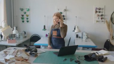 Orta yaşlı beyaz kadın deri atölye sahibi cep telefonuyla konuşuyor, kesme masasında deri ve süet parçalarını kontrol ediyor ve dizüstü bilgisayarda daktilo kullanıyor.