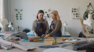 İki beyaz kadın deri atölye çalışanı dizüstü bilgisayarla masayı kesiyor, deri ve süet parçalarına bakıyor ve yeni ürün tasarımlarını tartışıyorlar.