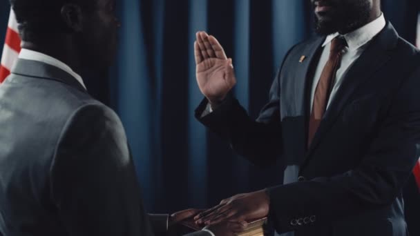 スーツ ネクタイ グラスに聖書やクルアーンで誓いを与える ひげを被った中年男性政治家の中年のタイルのショット 背景に手を挙げたアメリカの旗とダークブルーのドレープ — ストック動画