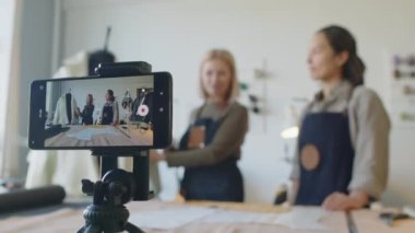 Tripodla akıllı telefonun yakın plan seçmeli görüntüsü ve arka planda bulanık önlüklü iki beyaz kadın atölyede duruyor ve çevrimiçi moda kursu için dikiş dersleri kaydediyorlar.