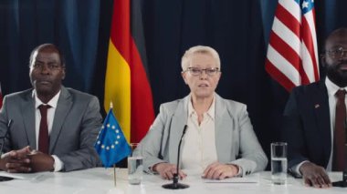 Orta yaşlı beyaz kadın başbakanın uluslararası sempozyumda erkek Afrikalı Amerikalı meslektaşlarıyla, İngiltere, AB, ABD, Alman bayrakları ve parlayan basın kameralarıyla konuşurken orta ölçekli bir fotoğrafı.