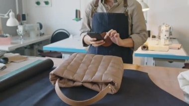 Genç bir kadın deri atölyesinin önlüklü orta yakın çekimi deri üzerinde yeni el çantasıyla masanın yanında duruyor ve sosyal medyada satışa sunulan fotoğraflarla güncelleme yapıyor.