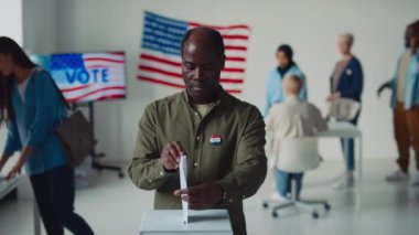Orta yaşlı, haki yeşili tişörtlü Afro-Amerikan bir adamın devlet referandumunda seçim merkezinde oy kullanırken, kameraya bakarken ve gülümserken orta yavaş portresi, arka planda ABD amblemi