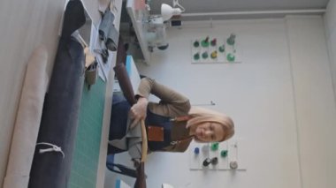 Orta yaşlı, beyaz önlüklü bir kadının atölyedeki masaya doğru yürüdüğü, deri rulolarla uzandığı ve süet parçalarını düzleştirdiği dikey bir fotoğraf.