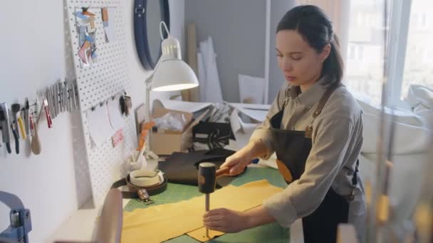 エプロンの若い女性職人ワークショップ従業員のミディアムショット金属パンチツールとハンマーで革の詳細を作る — ストック動画