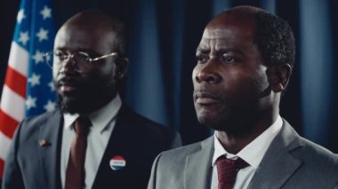 Afrika kökenli Amerikalı siyasi parti liderinin kötü seçim sonuçlarını duyması, tokalaşması, hayal kırıklığıyla başını sallaması ve arka planda ABD bayrağıyla onu rahatlatan siyahi erkek milletvekilinin yakın plan çekimi.