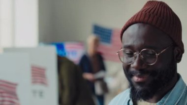 Kırmızı bereli, gözlüklü, gülümseyen ve kameraya poz veren orta yaşlı Afro-Amerikan bir adamın ve bulanık arka planda seçim bölgesinde oy veren farklı insanların yakın çekimde odak portresi.