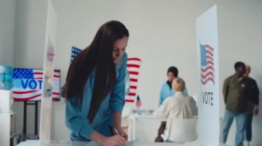 Eyalet referandumu sırasında sandık merkezindeki seçim kabininden orta ölçekli bir çekim, çeşitli erkek ve kadınların yürümesi, oy pusulasına yazı yazması, sonra da sandığa atmak için ayrılması ve arka planda ABD bayrakları