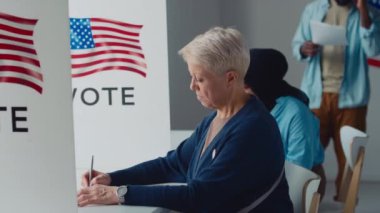 Orta yaşlı beyaz kadın ve Müslüman kadının oy kullanma merkezindeki kabinlerde otururken, oy pusulasında yazı yazarken ve ayrılırken, çeşitli Amerikan vatandaşlarının kuyruğa girerken, arka planda ABD bayrağı amblemi