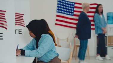 Siyahi İslami tesettürlü genç bir kadının oy pusulasında oturduğu ve oy pusulasını doldurduğu ve tanınmayan kadınların ABD bayrağının yanındaki bulanık arka planda sohbet ettiği orta boy bir fotoğraf.